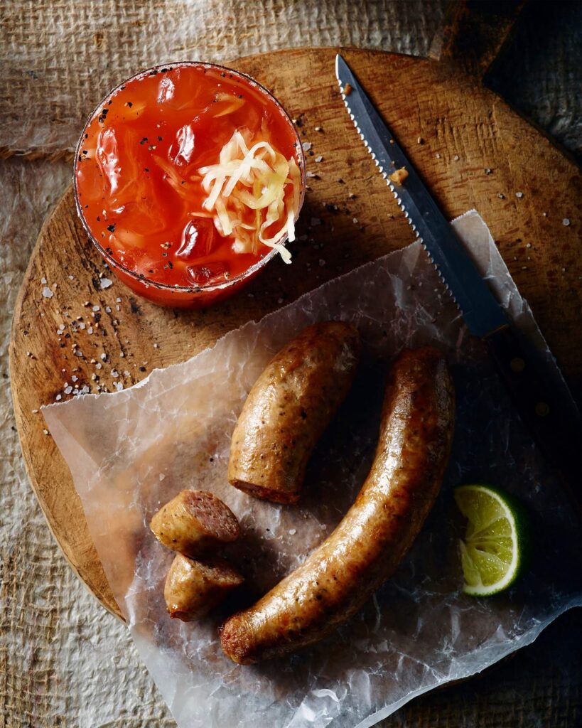 image of caesar with sauerkraut as garnish, knife resting beside and cooked sausage on cutting board