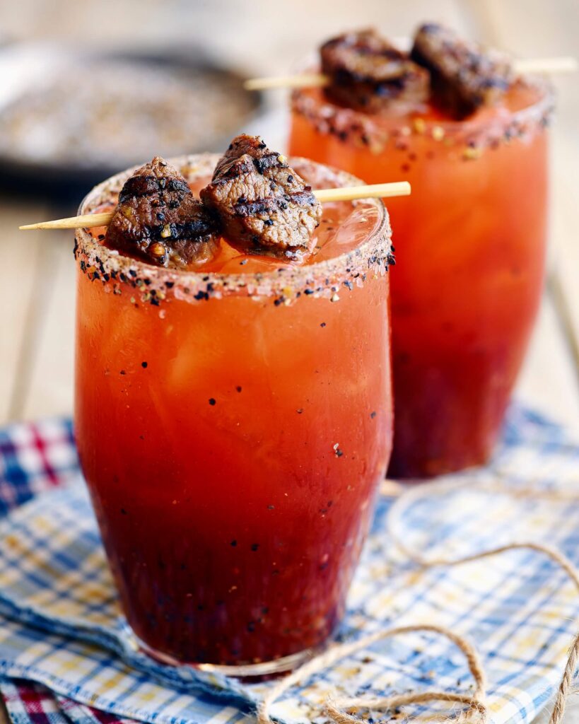 two Caesar drinks with beef skewer garnish resting on top of table cloth
