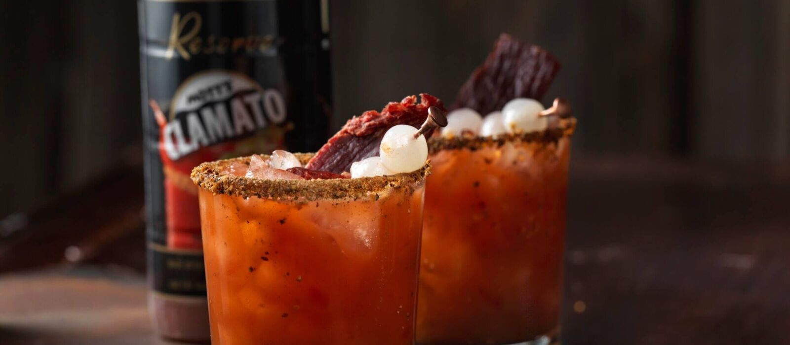 Two caesar's garnished with pepperoni sticks and pickled inions in front of Mott's Clamato reserver bottle